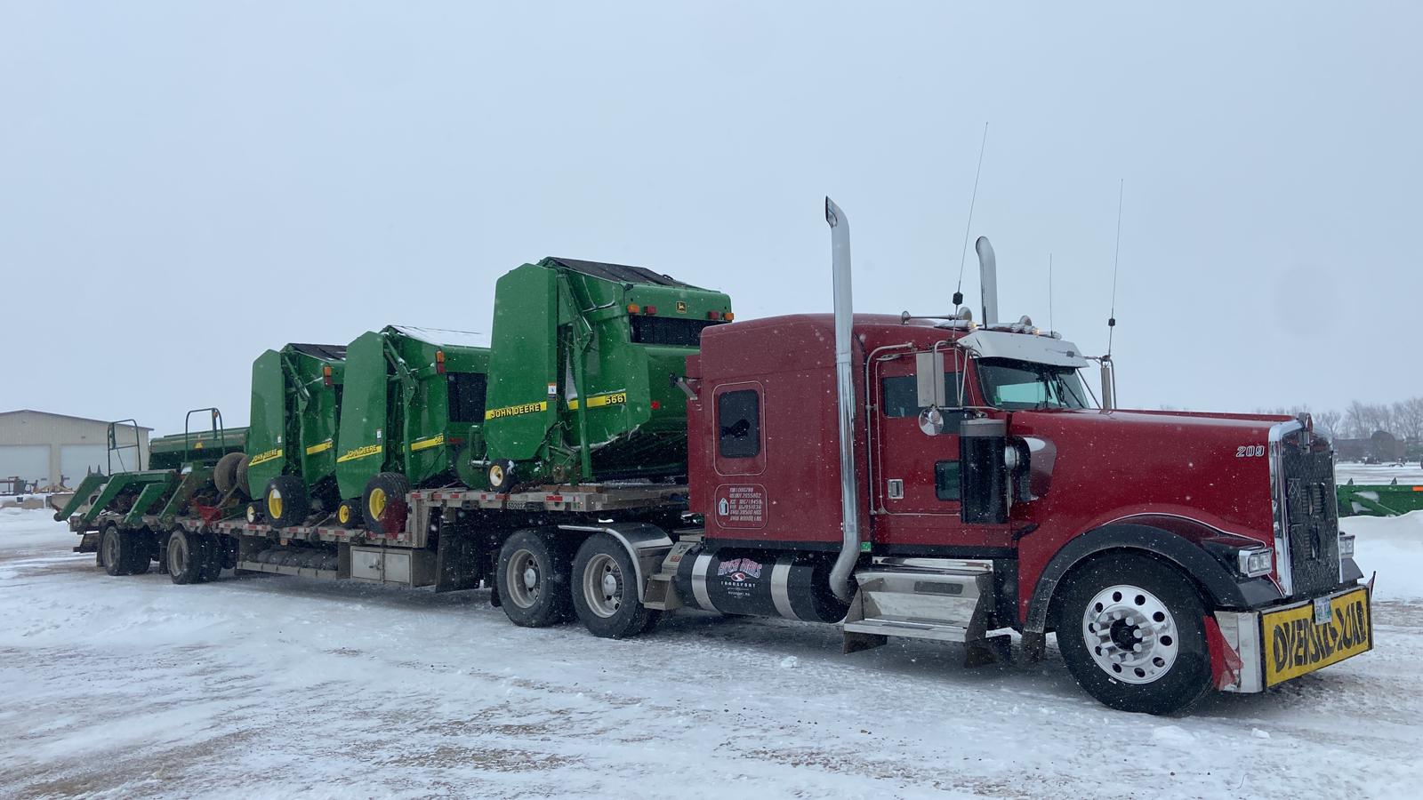 Red Kenworth pulling a River Trails Transport tandem step deck loaded with John Deere round balers